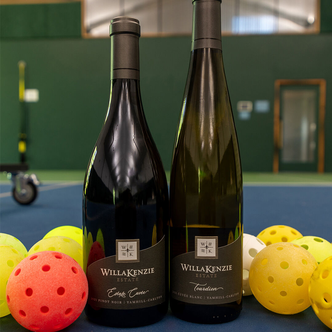 Two bottles of WillaKenzie wines surrounded by pickle balls on pickle ball court.